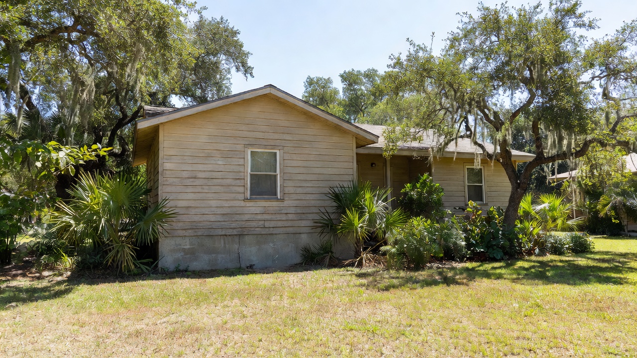 Single-story wooden Florida home with concrete foundation, windows, and surrounding trees and vegetation under bright daylight