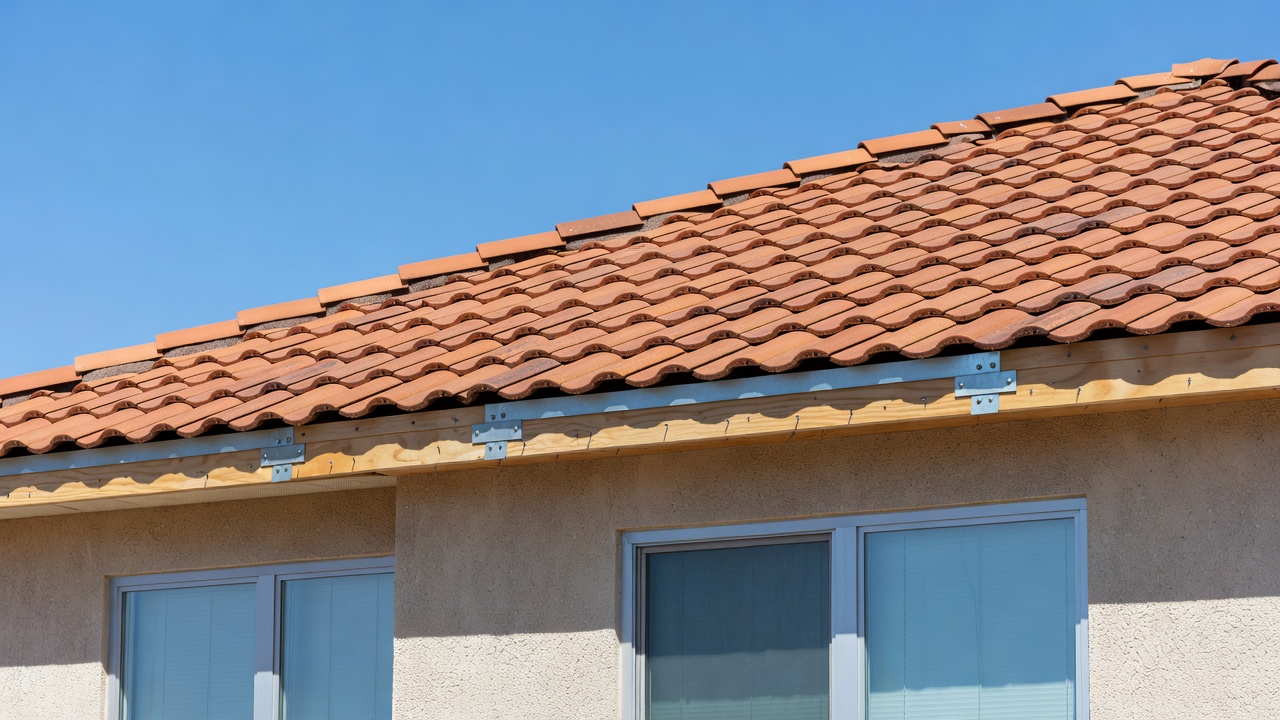 Close-up of red-tiled roof with metal hurricane straps, exposed roof deck nailing, and impact windows on stucco exterior under clear blue sky